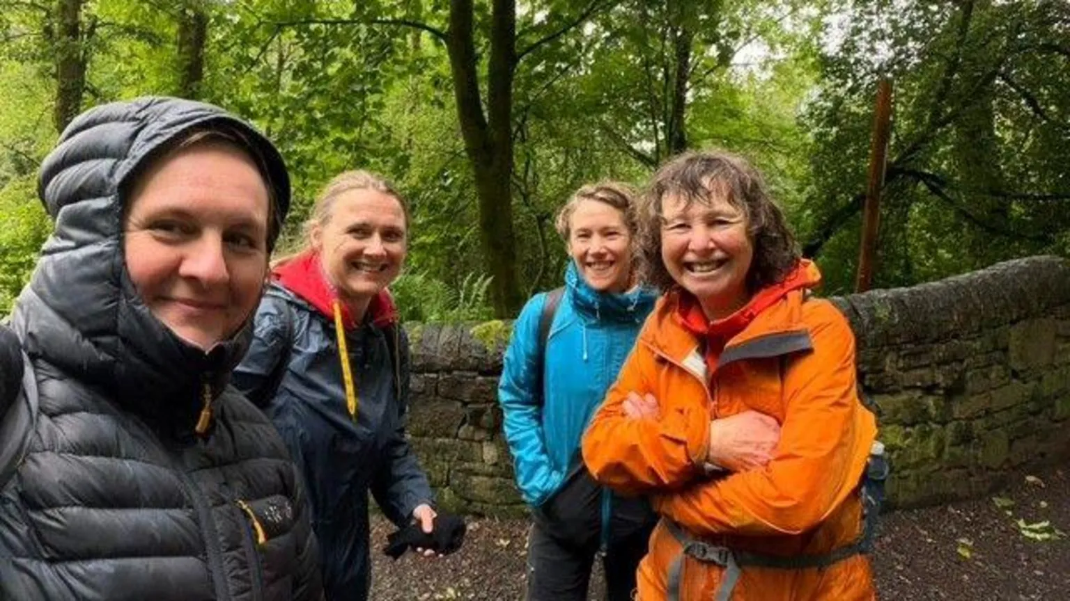 Lorraine McCall Four women are standing in front of a stone wall in a forest. They are wearing waterproof jackets and it appears to be raining but they all have big smiles. 