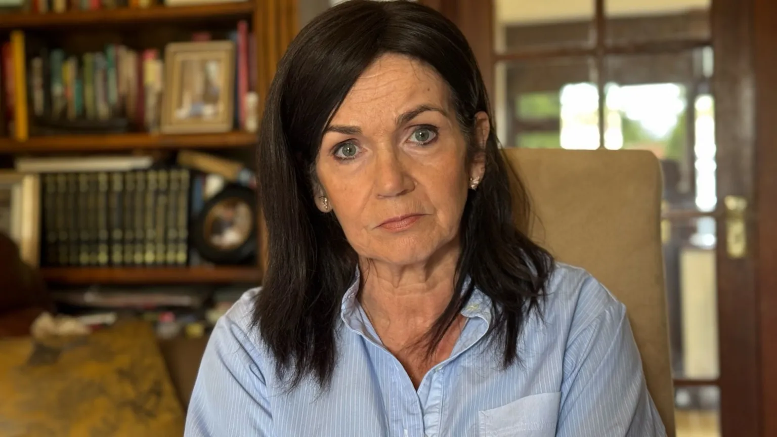 A woman with dark shoulder-length hair and a blue shirt. There are shelves in the background with books and photographs