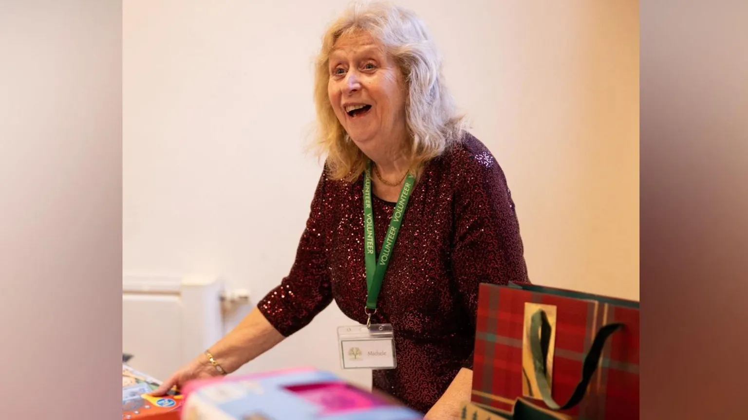 Allwyn A woman wearing a sparkly dress and green lanyard smiles while looking away from the camera. She is surrounded by gift bags and boxes of toys.