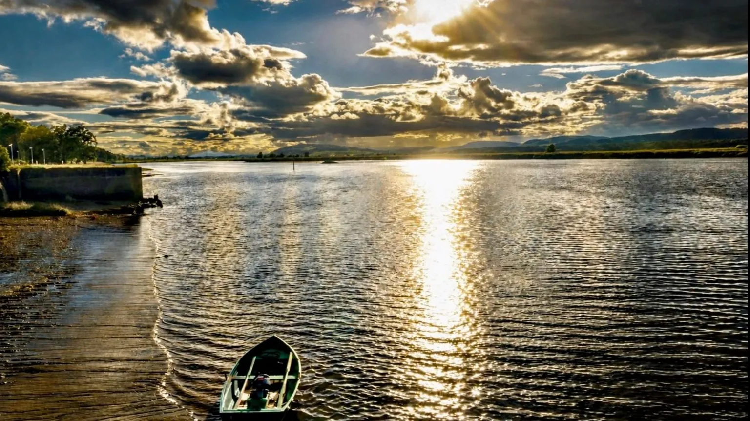 Michael O'Kane Rays of sunshine hit the water despite mostly dark clouds in the sky. A small rowing boat is sitting in the bay.
