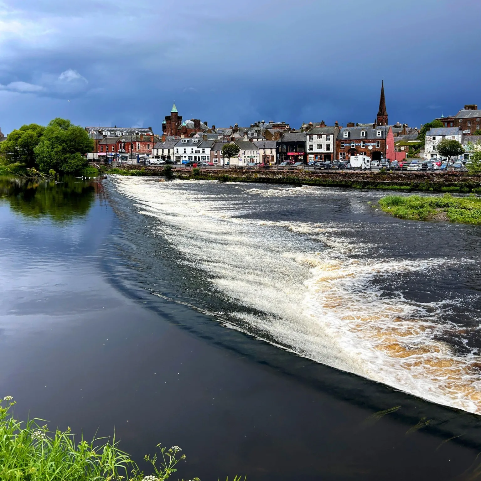 Glyn Booton A view of a river at Dumfries, with the town in the distance and part of the sky overcast and the other half sunny