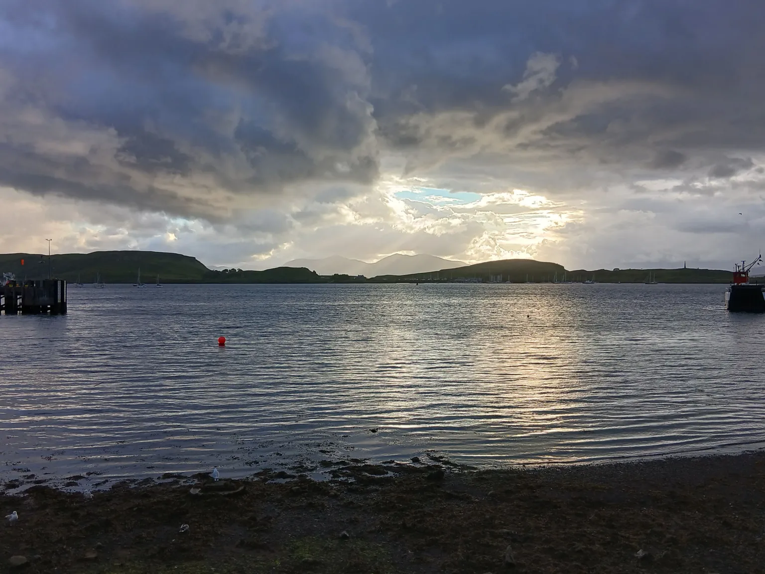 Heather Black Oban, with the sky mixing blue sky with very dark clouds. The view is from across water with the island in the distance.