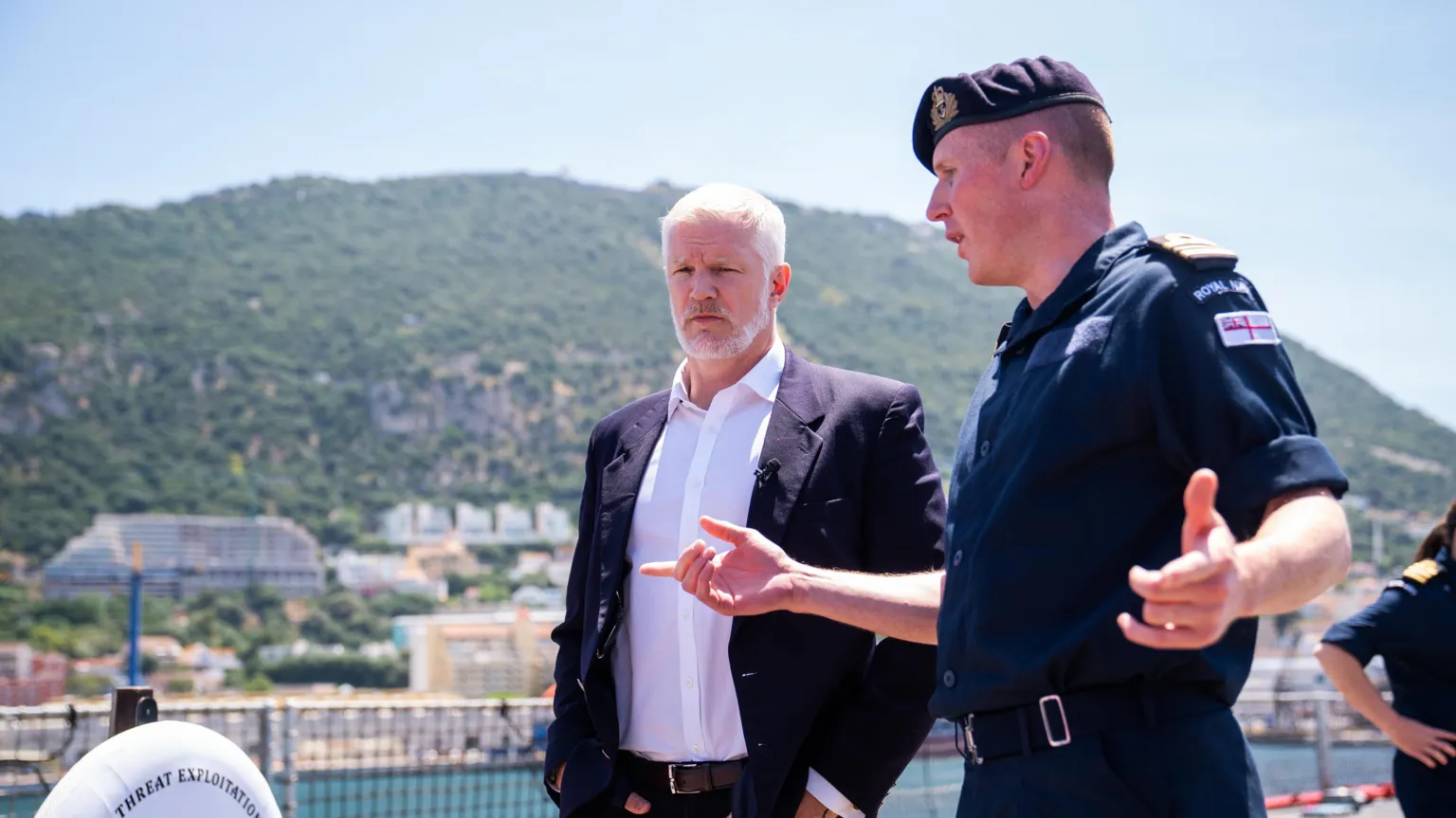 Two men, on the left is Al Carns wearing a dark suit jacket, trousers and brown belt, with a white shirt underneath. To his right is a man wearing a military uniform. They are pictured talking on an outside deck of a boat. In the distance, behind them there is a built up residential along the water and a mountain.
