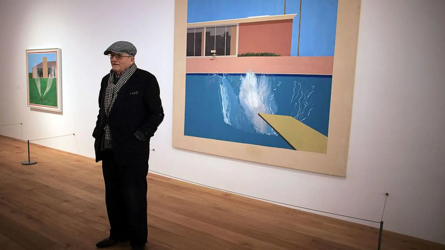 Christopher Furlong/ David Hockney stands in dark clothing in front of 'A bigger splash 1967', one of his most famous paintings