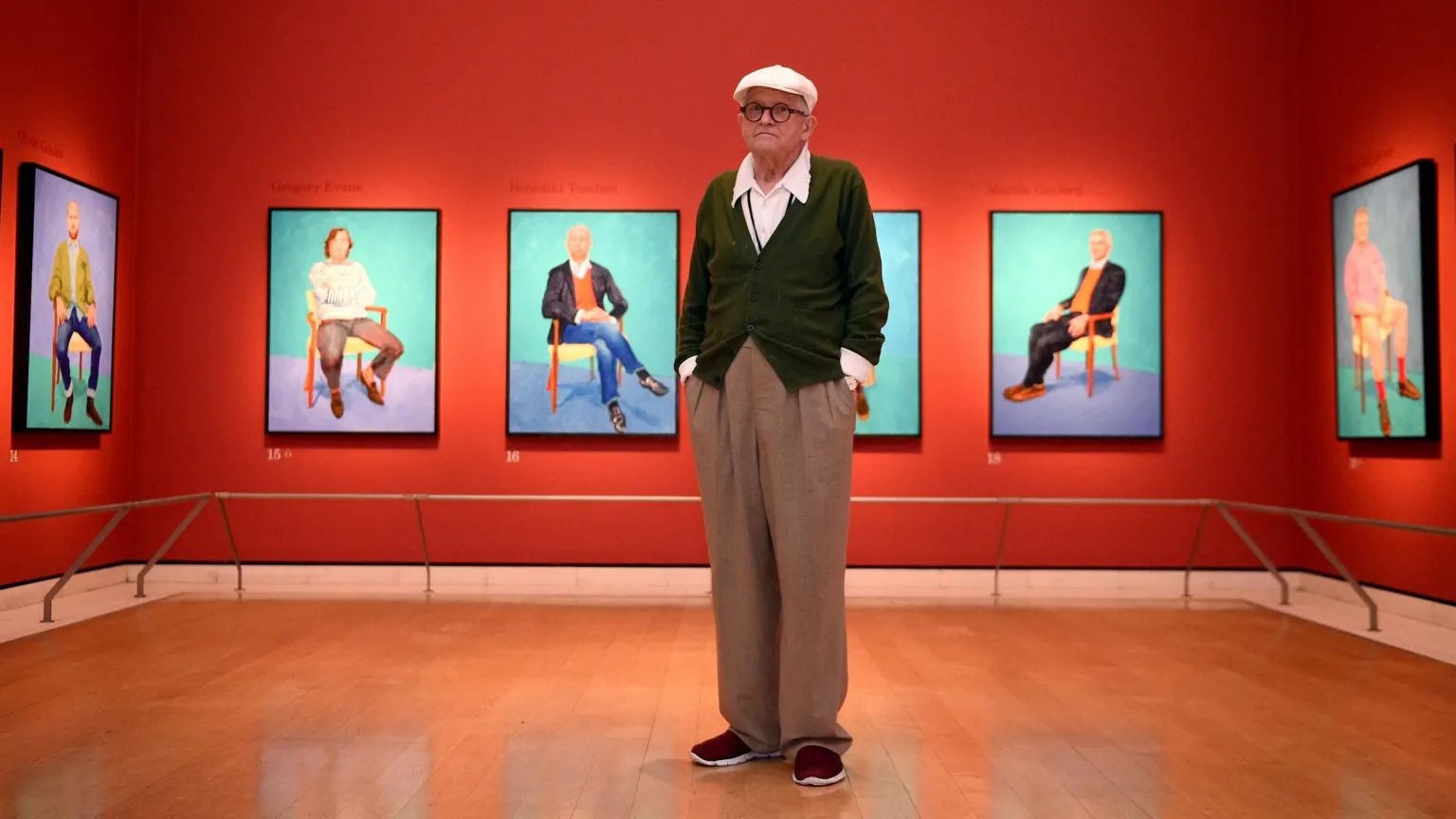 David Hockney stands in brown trousers, a green cardigan in a gallery. His 'still-life' portraits are hung on the orange walls behind him.