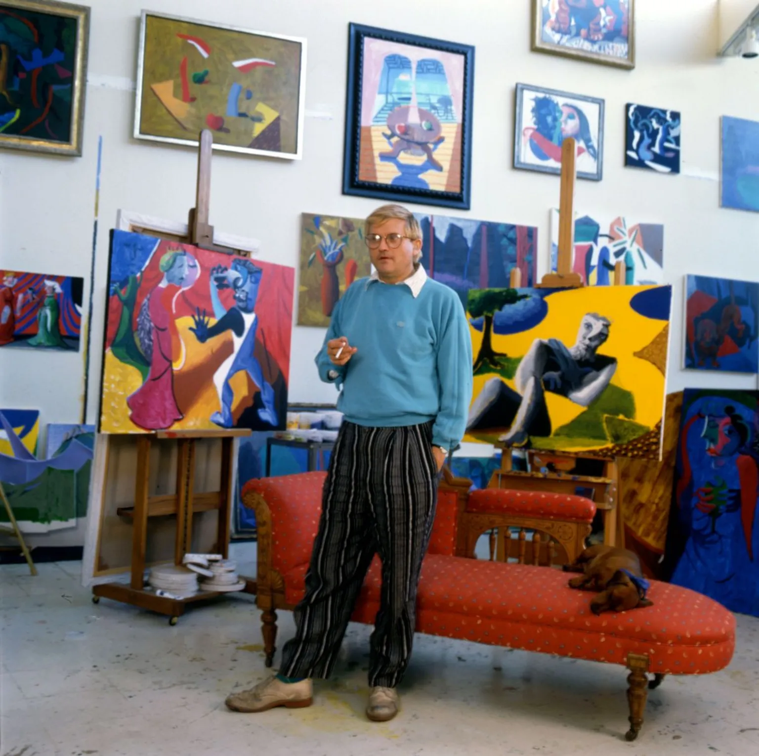 Portrait of English painter David Hockney, dressed in a light blue sweatshirt and tan trousers, as he stands in front of a chaise longue in his home, Los Angeles, California, 1987.