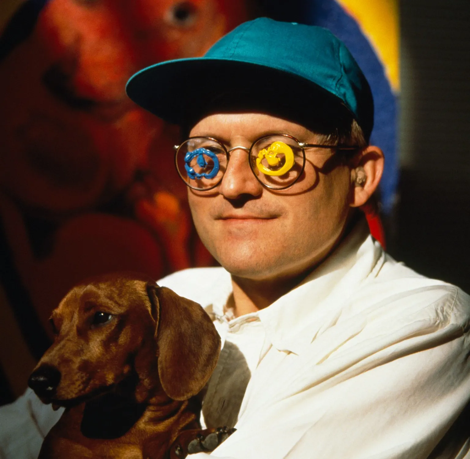 David Hockney is holding a brown dog and is wearing a blueish green baseball cap and glasses with spiral designs on them.