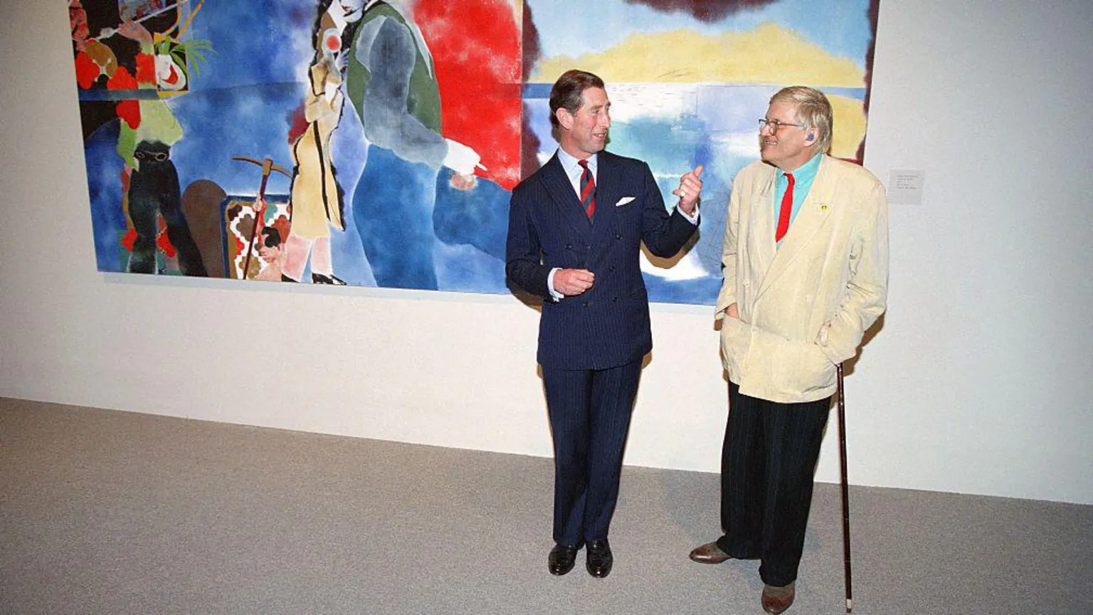 AFP via David Hockney next to King Charles in 1994. Charles wears a dark suit, while Hockney is dressed in a white blazer, red tie and blue shirt, with black trousers. Behind them is a colourful piece of art