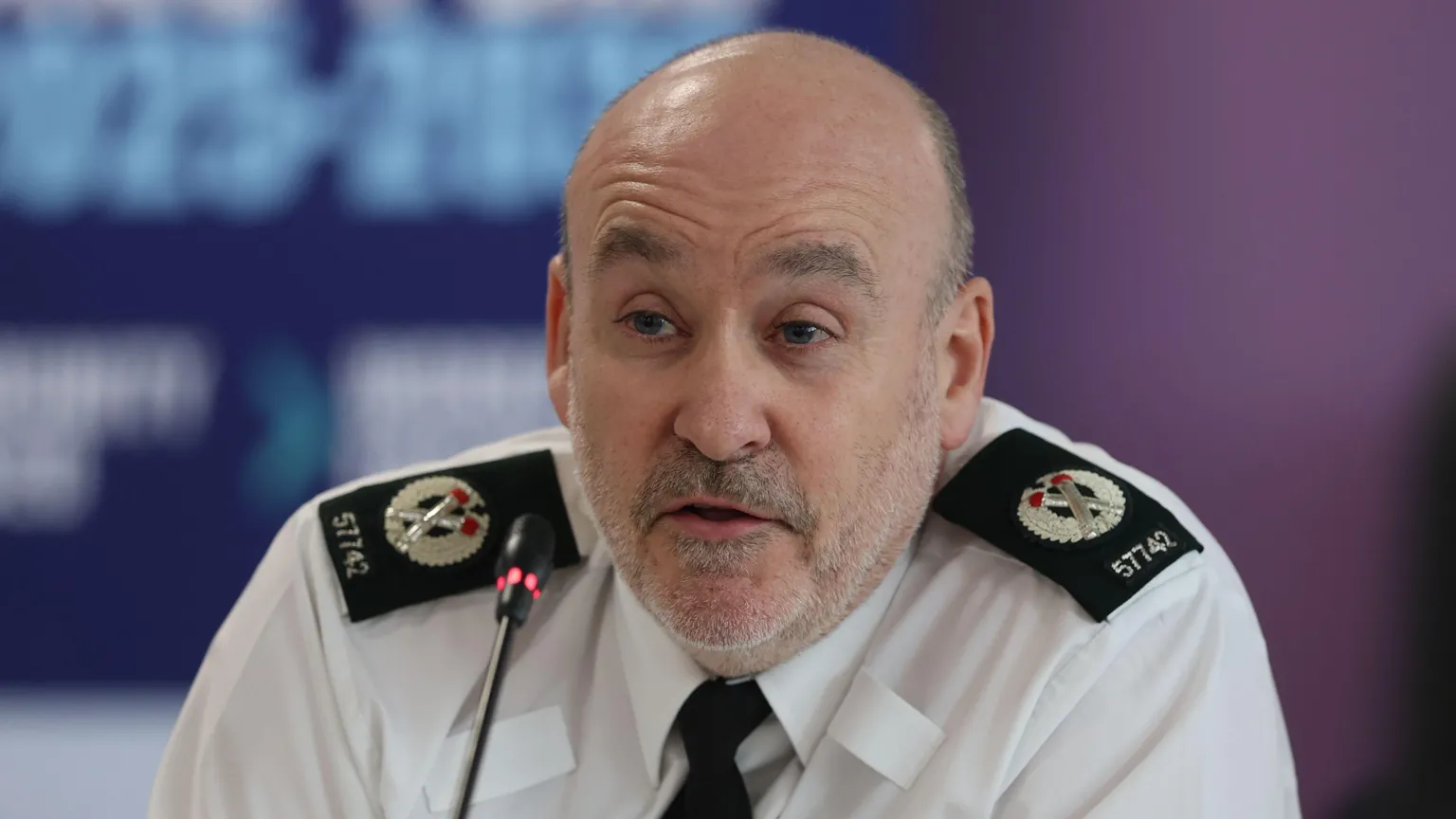 Ryan Henderson is pictured during a press conference. He has very short grey hair and a beard and is dress in a white uniform shirt.