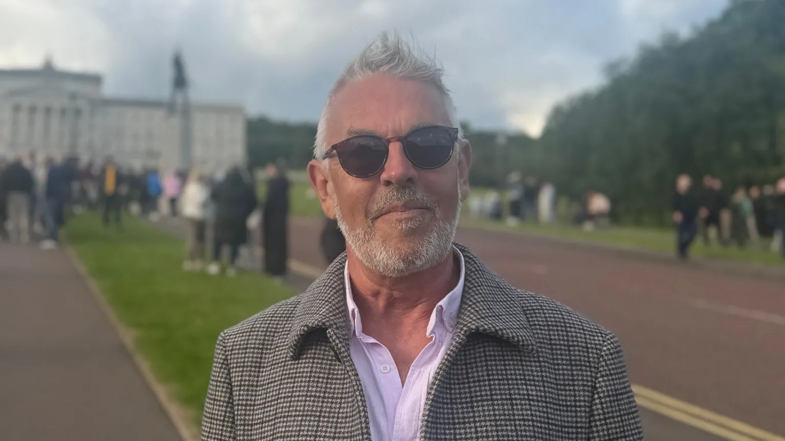 Paul corry is pictured in the Stormont Estate. He has short white hair and a beard and is facing the camera wearing sunglasses. Behind him is the Stormont building in the distance and a large number of people, all are out of focus.