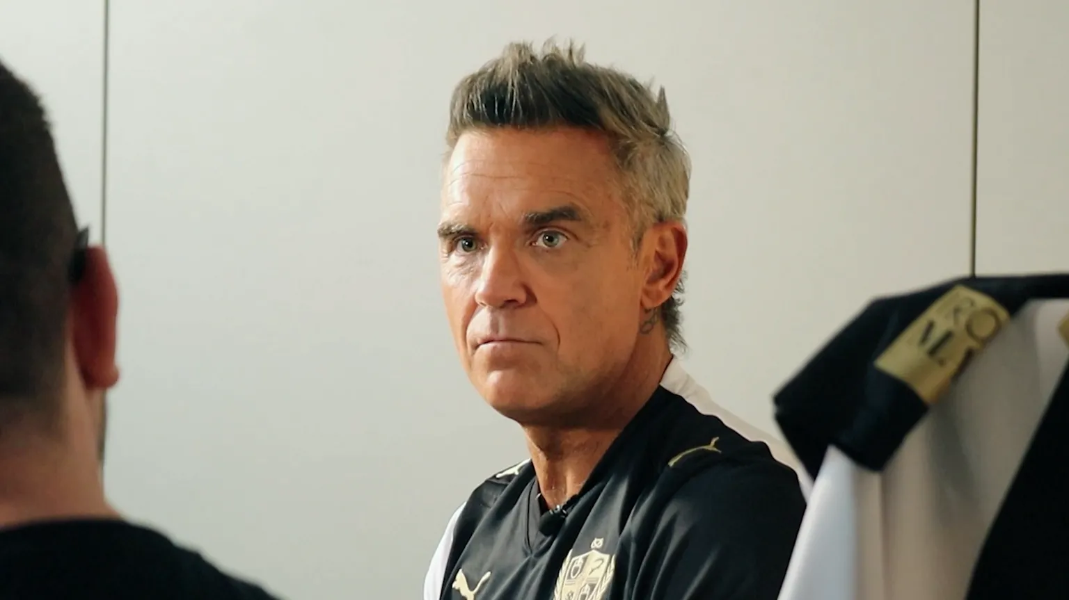 Port Vale Football Club Robbie Williams, a man with grew spiked-up hair, is looking at a man who has his back to the camera to the left of the shot. He is wearing a black and gold football shirt.
