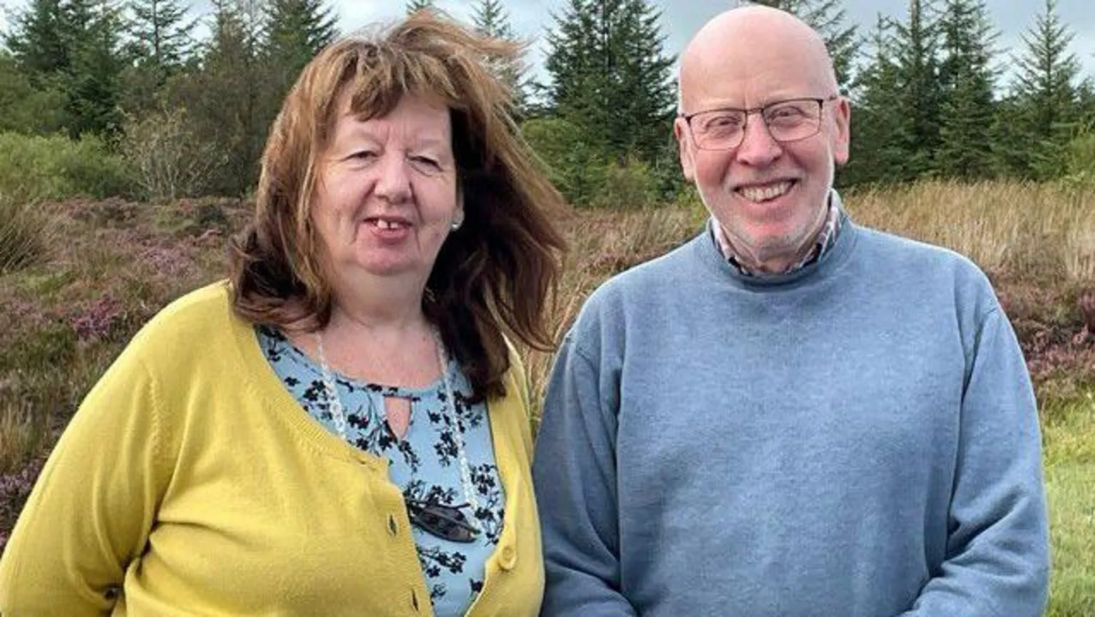 WAVE Trauma Centre/PA Wire A woman with brown hair, a blue top and yellow cardigan stands beside a man in a blue jumper. He wears glasses. They are outside. Green fir trees are visible behind them.