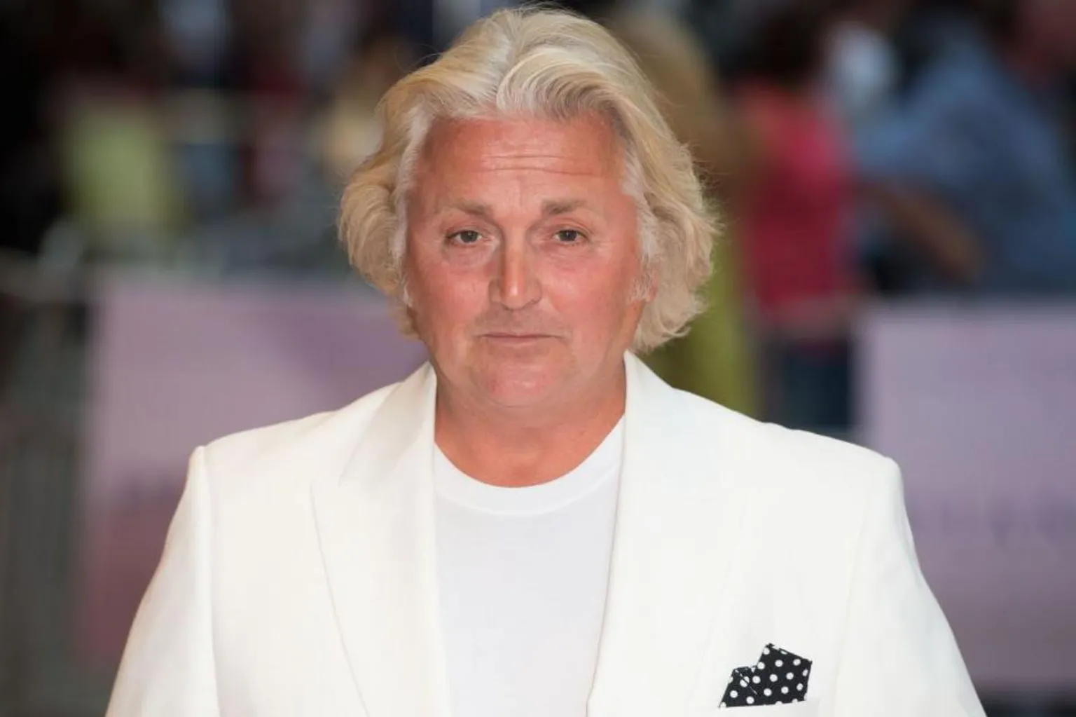 Head and shoulders shot of David Emanuel. He has white hair and looks at the camera. He is wearing a white t-shirt and white shirt with a black and white polka-dot scarf in the left-hand pocket.