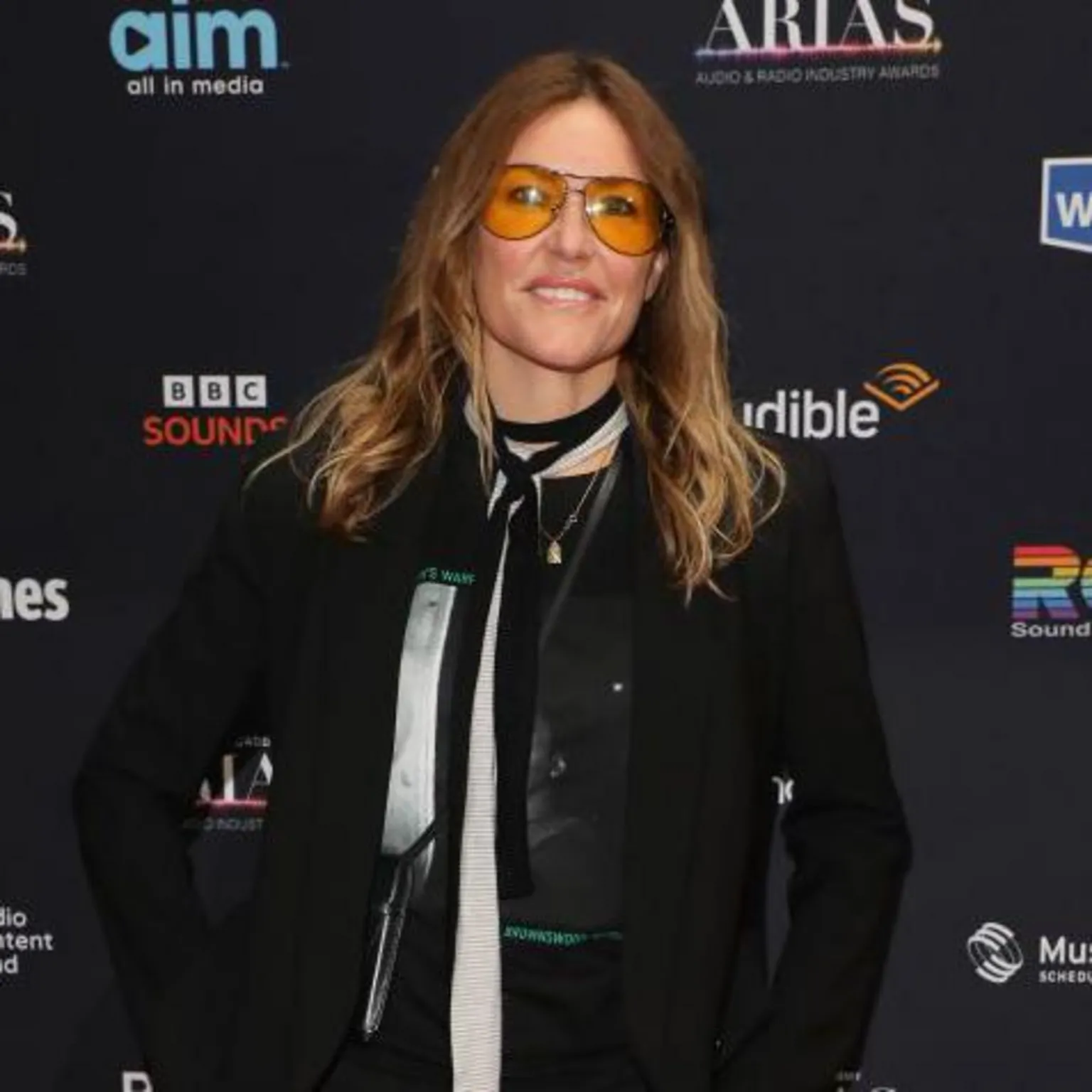 Cerys Matthews wearing yellow sunglasses. She has long blonde hair and bits her lower lip as she smiles at the camera. She is wearing a black blazer, with a black and white tie.