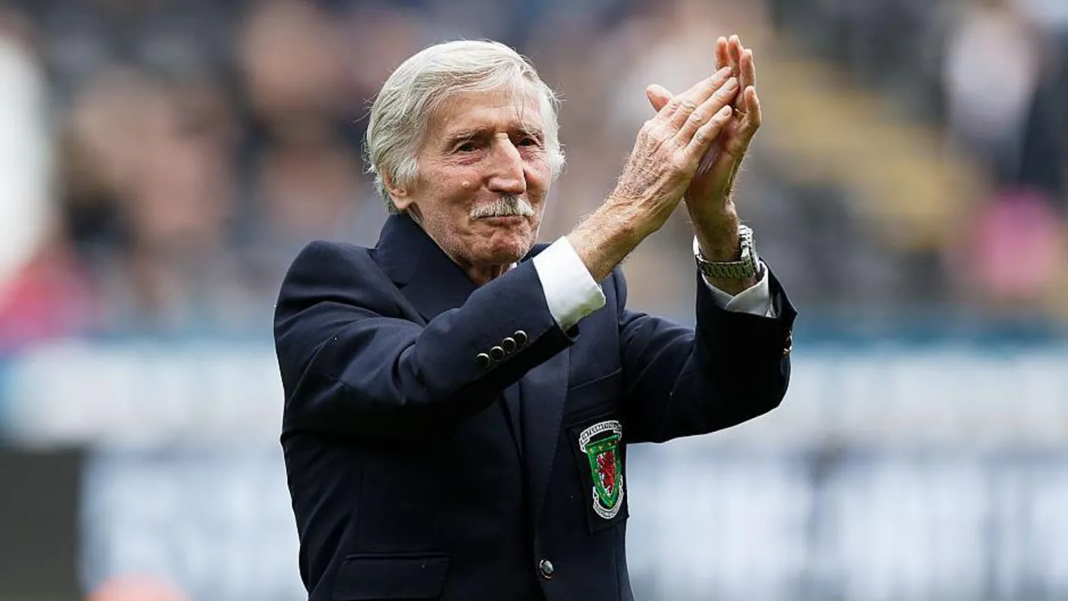 Cliff, who has white short hair and a moustache, has his arms raised clapping. He has a blue blazer on with the Wales logo.