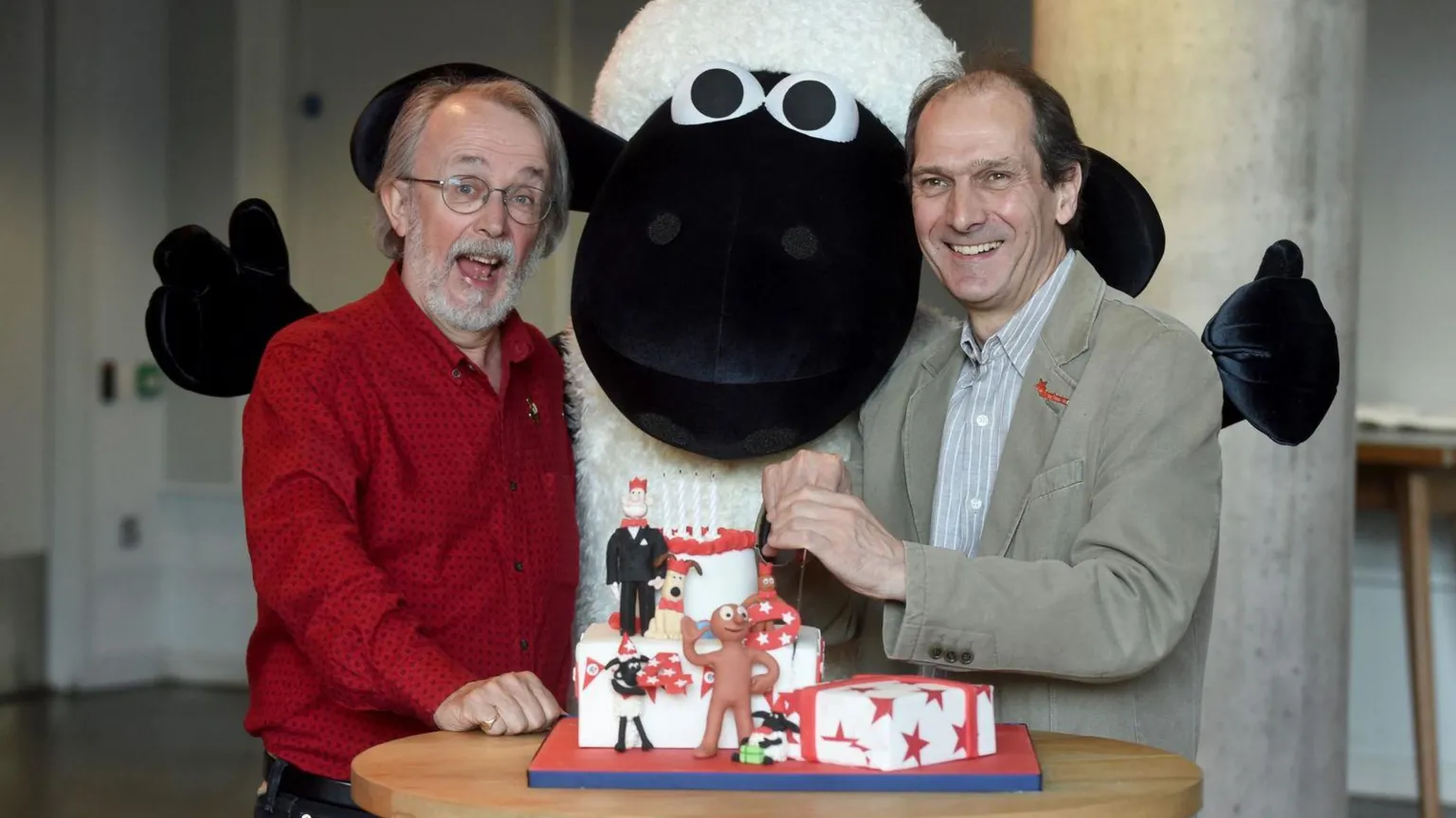 Decorated cake on a small round table in the foreground, topped with figurines and red star patterns, with a knife cutting into it. Behind the table stands a large Shaun the Sheep mascot with a black face and white wool, flanked by Lord and Sproxton who are wearing long-sleeved shirts. 