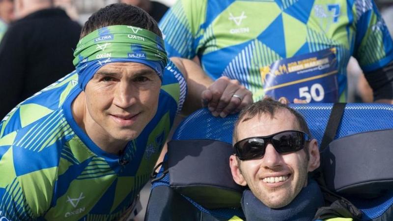 Kevin Sinfield (left), and Rob Burrow, smiling together at the start line of a race, with Sinfield wearing a green and blue running top and bandana. Rob Burrow is wearing sunglasses and in a wheelchair. 