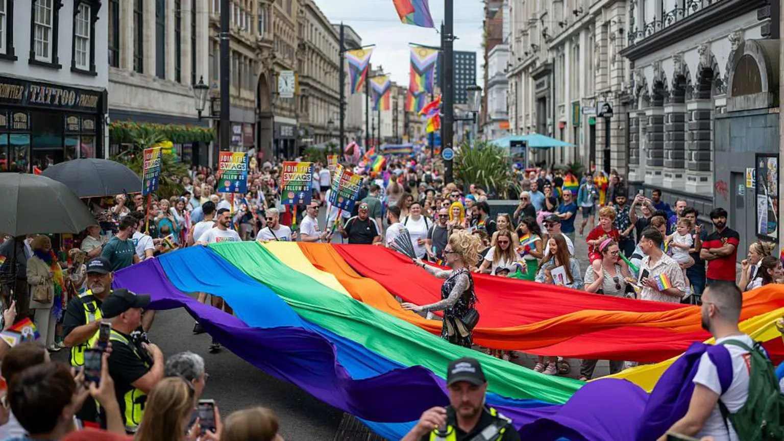 A wide image showing crowds walking in the Pride parade on Cardiff St Mary's Street. A large pride rainbow flag is being carried by the crowd.