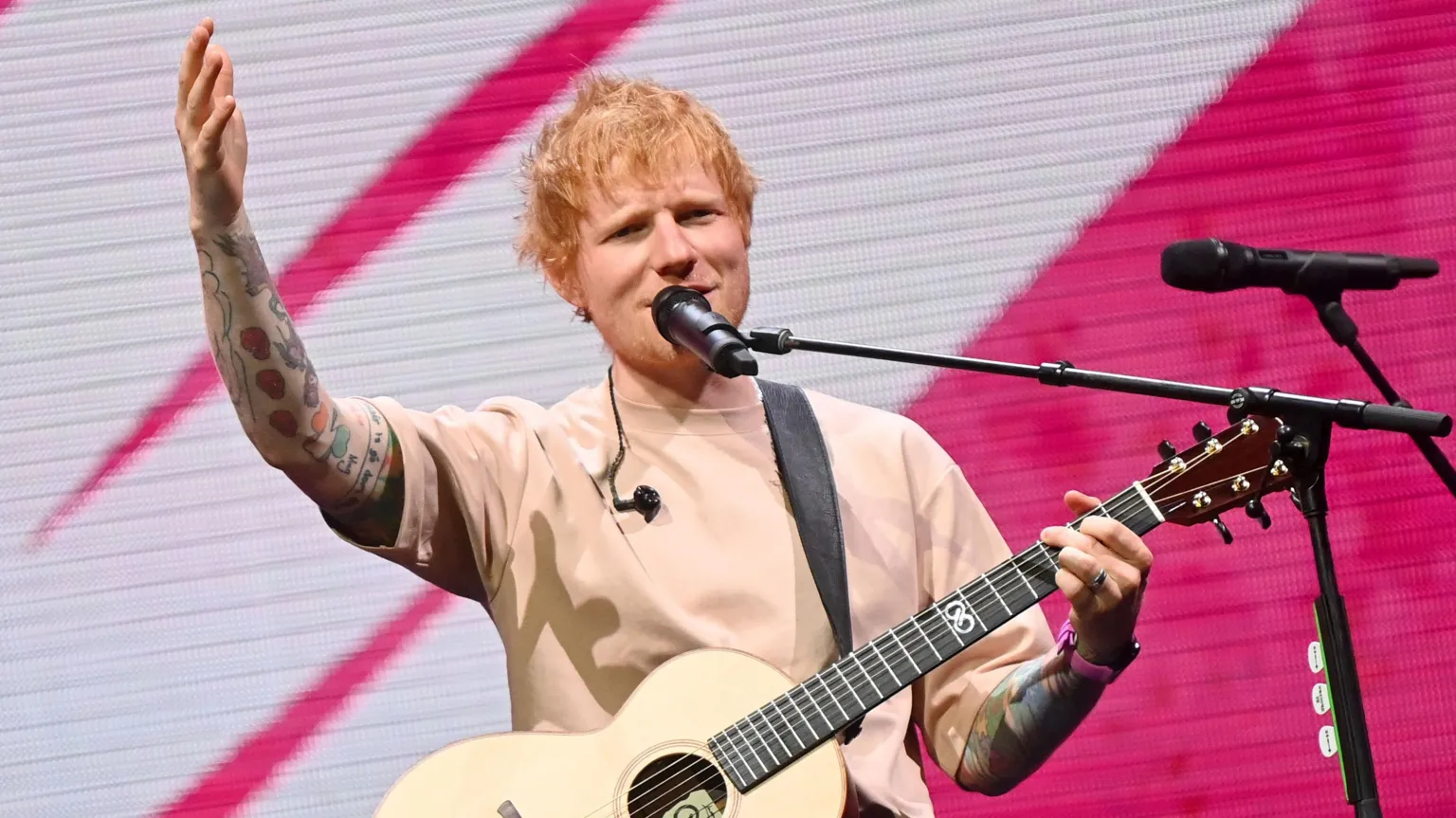 Ed Sheeran singing into a microphone on stage with one hand on the neck of his guitar and the other raised.