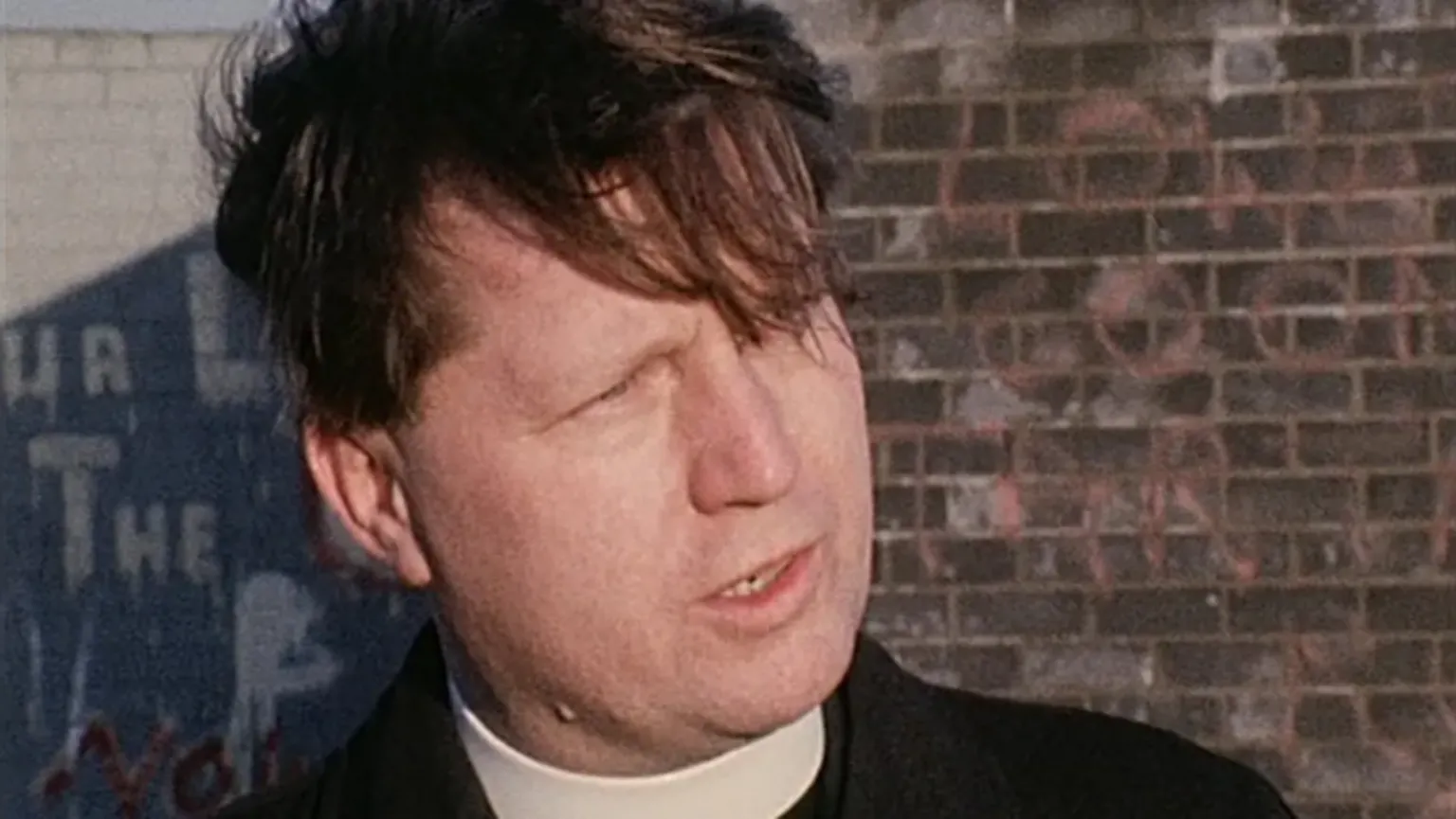 A man with dark, medium length hair is standing in front of a red bricked wall. His hair hangs down over his eyes as he looks to the right of the camera. The man is wearing a black shirt and coat with a clerical collar.