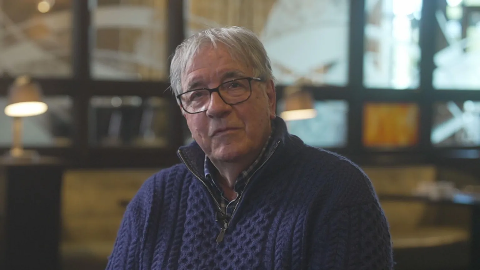 A man with grey hair is sat looking at the camera. He is wearing glasses and a thick, navy twead jumper with a quarter zip. Behind the man is an out of focus industrial space with lots of windows and open space. There are some lamps placed in front of the windows.