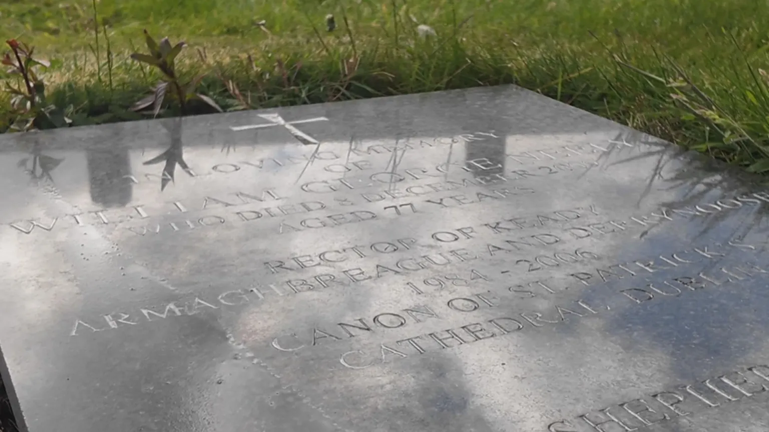 A large flat headstone lays on the ground, surrounded by grass which pokes over it's edges. It's a black marble like stone with a cross at the top. Only some text is visible, showing the name 'William George Neely' and his age, of 77. Below it reads 'Rector of Keady'.