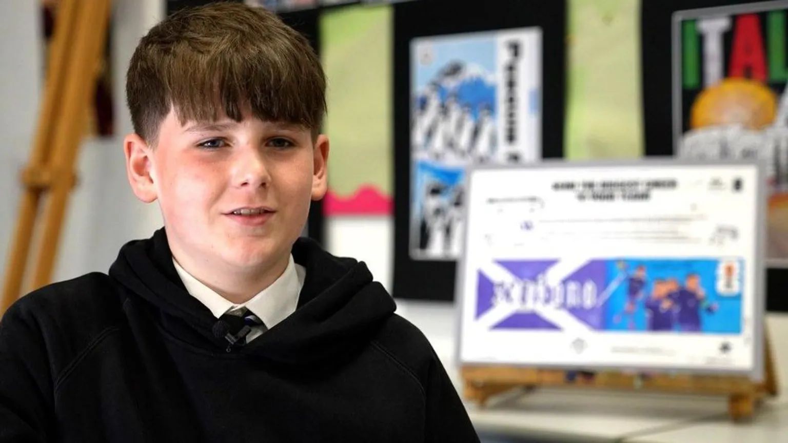 A boy wearing a school uniform sitting beside a screen with a drawing on it