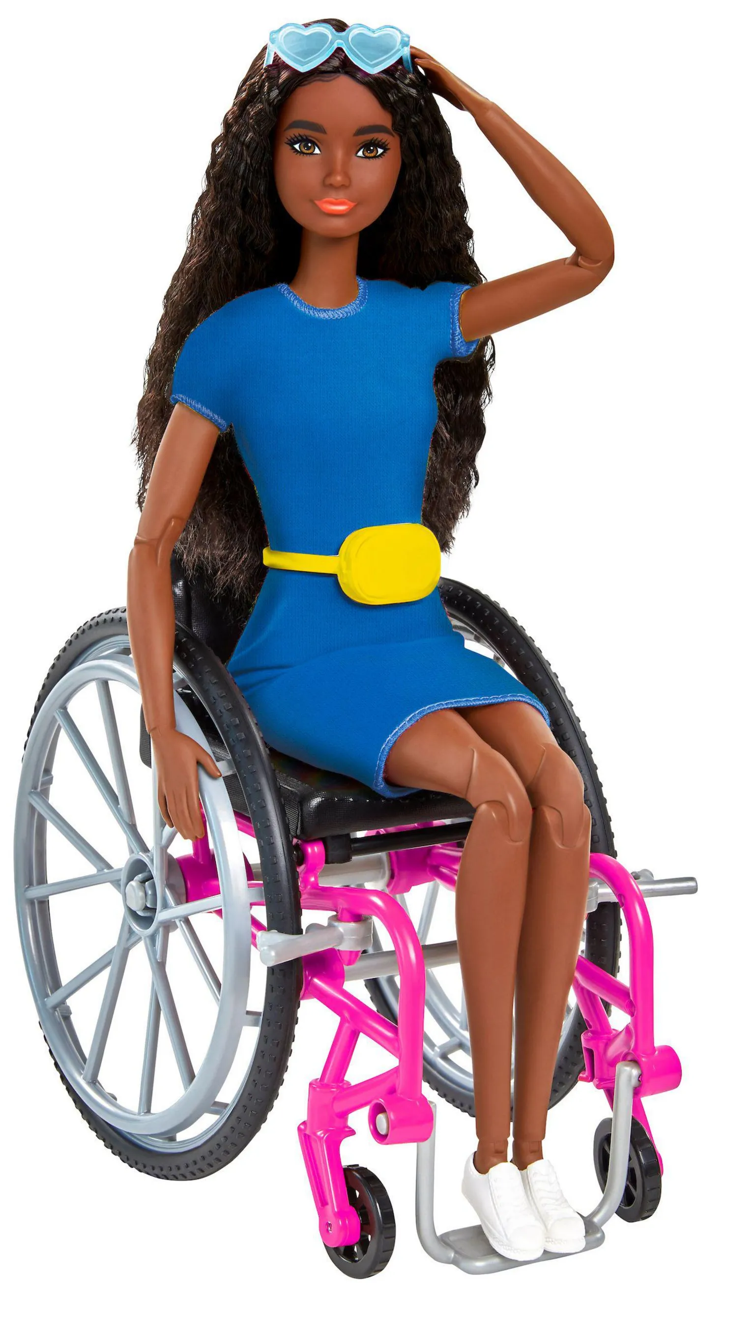 Mattell Inc Fashion doll seated in a pink-framed wheelchair with large grey wheels, wearing a blue dress with a yellow belt, white sneakers, and heart-shaped blue sunglasses on the head, against a plain light background.