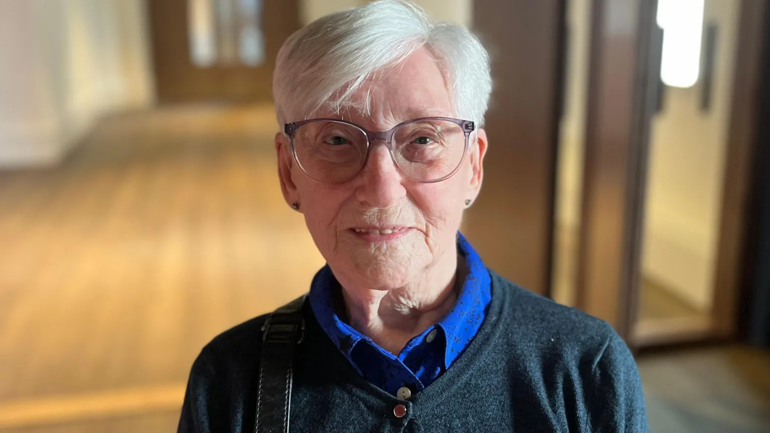 Close-up portrait of 90-year-old Mary with short white hair and glasses, wearing a dark sweater over a blue collared shirt, standing indoors with a softly blurred hallway background.