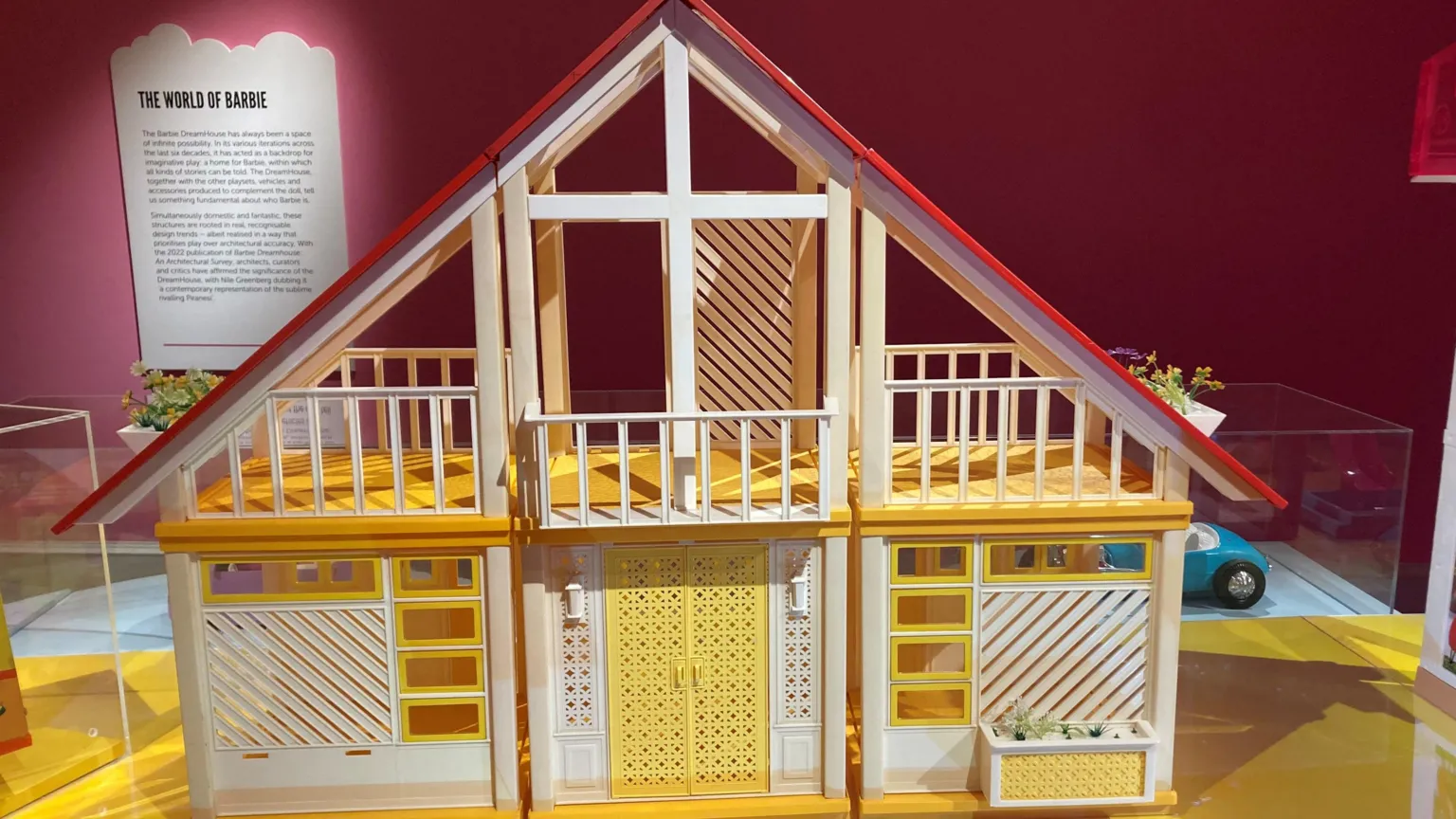 Model of a Barbie Dreamhouse displayed in a museum-style exhibit, featuring a two-level structure with white railings, yellow accents, and a red roof, set against a pink wall with an informational panel titled “The World of Barbie” and a small toy car visible in the background.