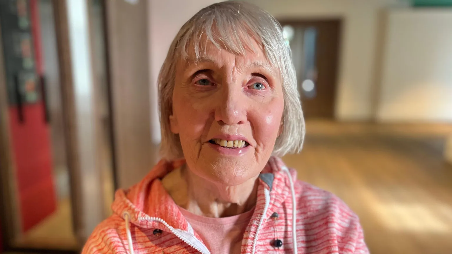 Close-up portrait of 90-year-old Margaret with short grey hair, wearing a pink zip-up hoodie over a light top, standing indoors with a softly blurred background.