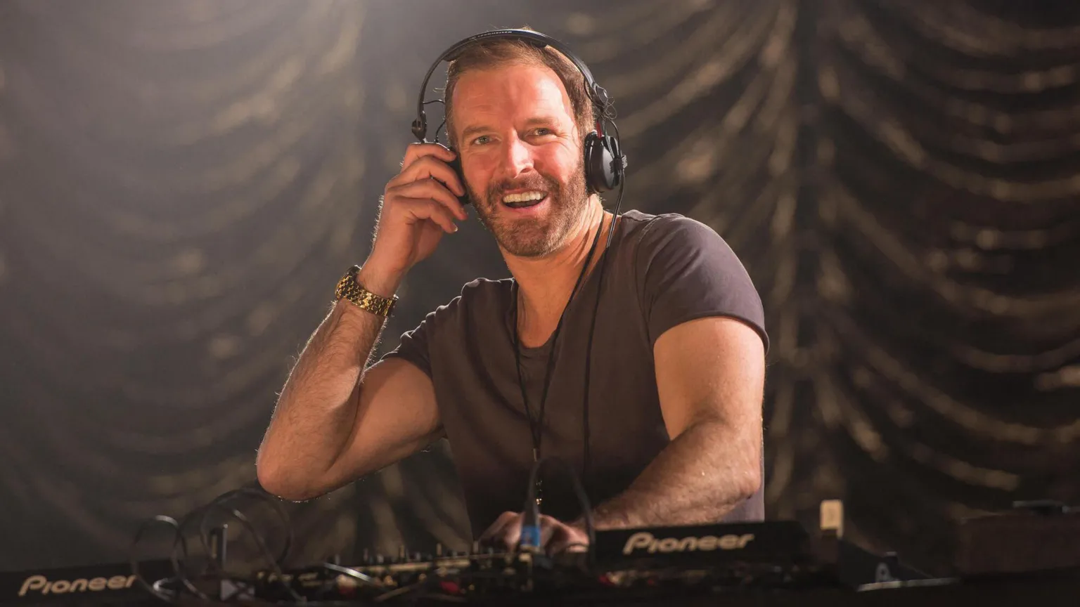 Alamy George Bowie DJs in front of a dark curtain. He has short brown hair and wears a dark-tshirt and gold watch