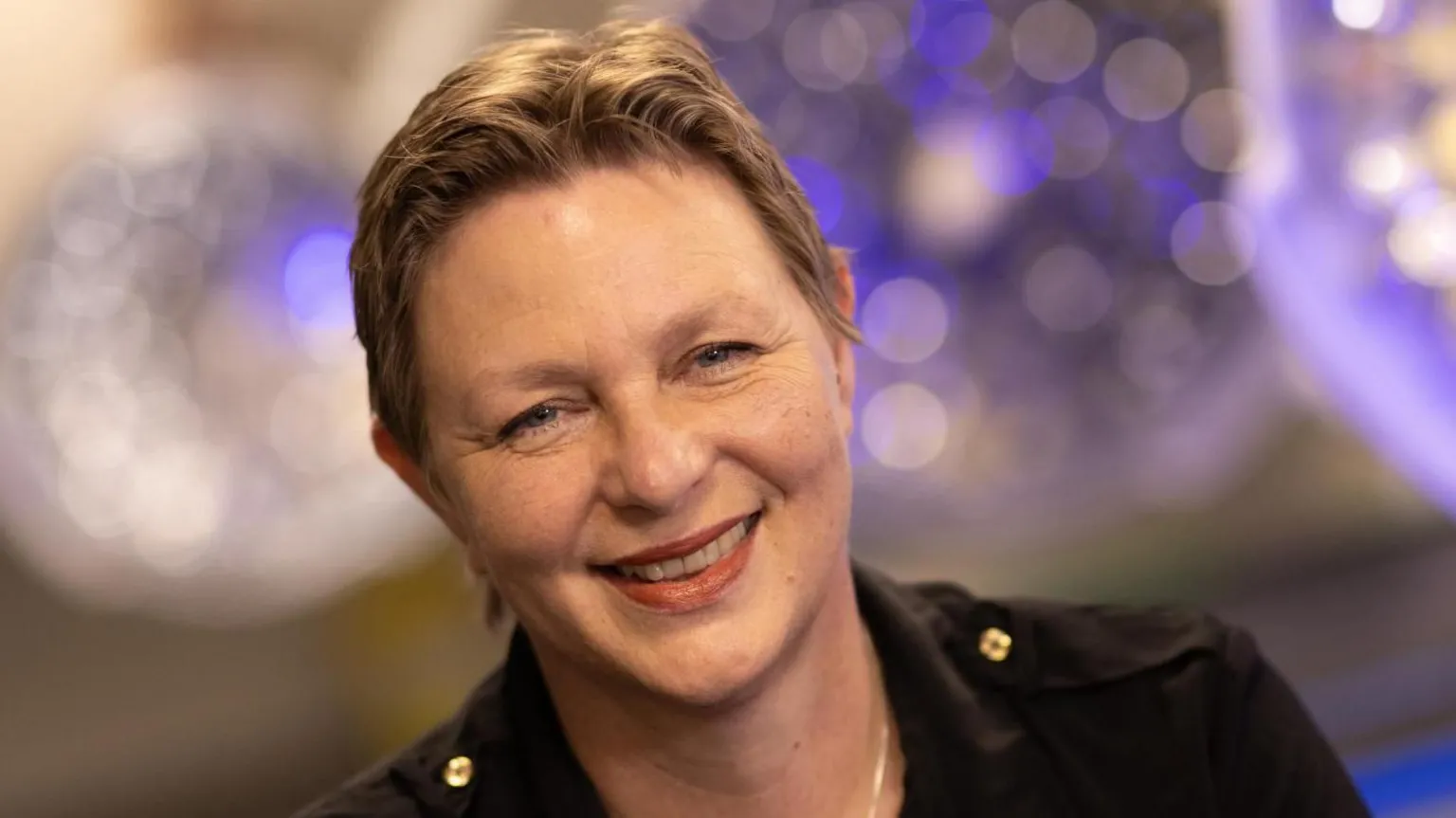  Dame Lucina Hackman is smiling widely at the camera. She has short, brown hair and is wearing a dark jacket. There are blurred lights behind her