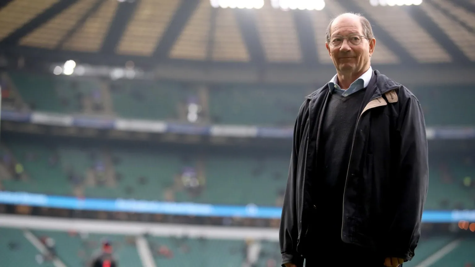  Ian Robertson is standing inside a rugby stadium which is blurred in the background. He has a bald head and glasses and is wearing a black jacket over a dark jumper and a light-coloured shirt.