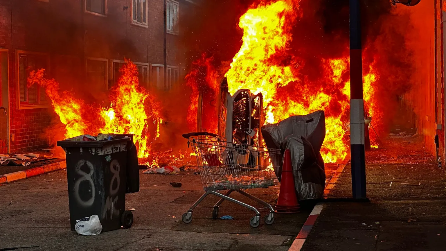 A wheelie bin and a trolley and a car are all on fire on a street. The kerb stones are painted red, white and blue.