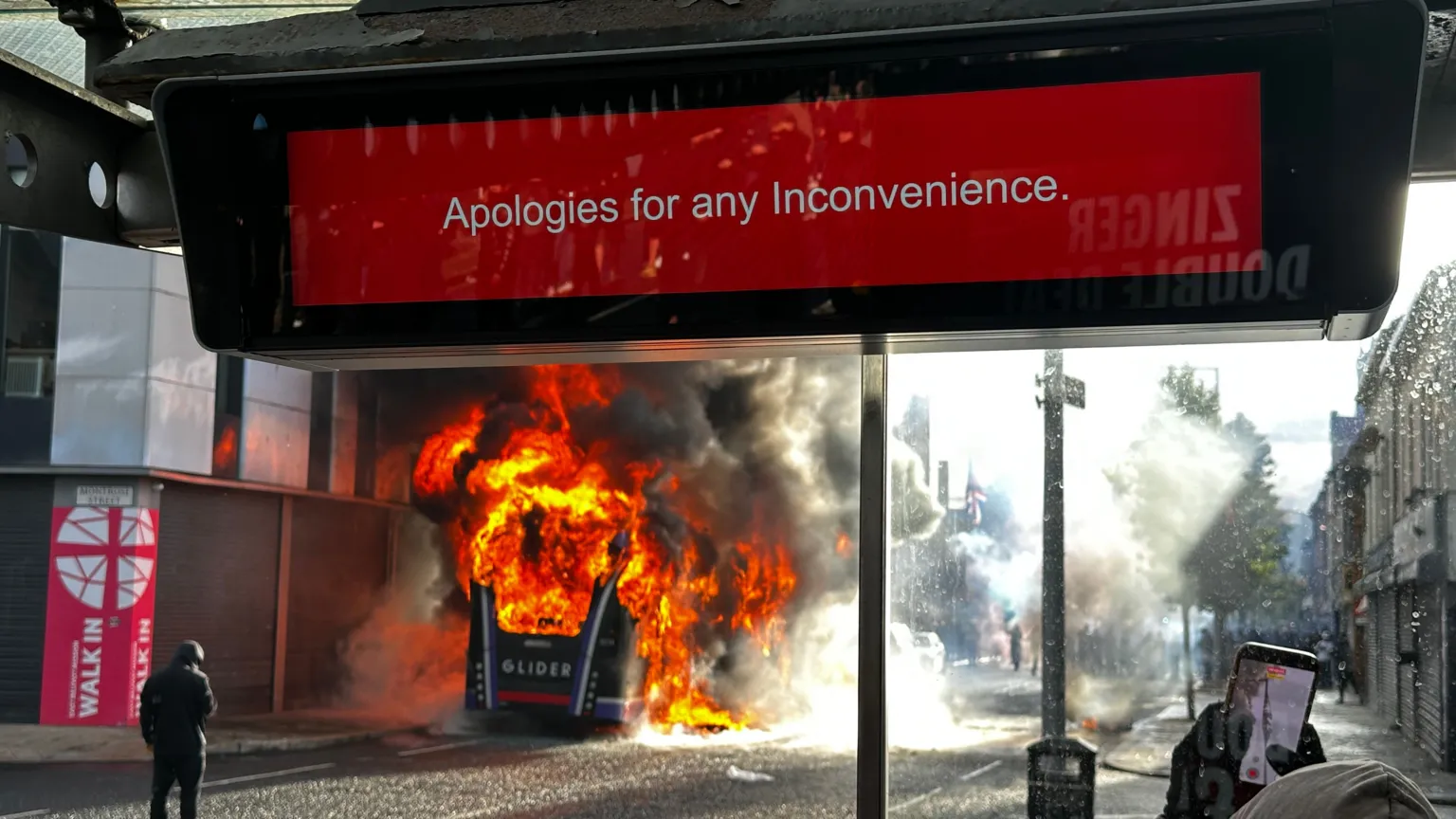A Glider bus is on fire on a street. There is a sign at a bus stop at the top of the picture which says - Apologies for any Inconvenience. At the bottom right hand corner, some is filming the bus on a mobile phone.