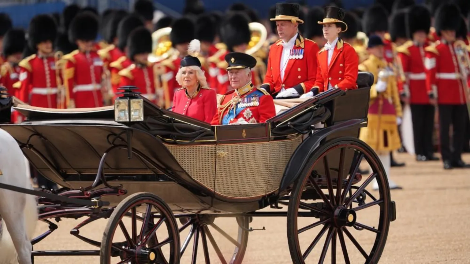 The King and Queen riding in a carriage. He is wearing the uniform of the Grenadier Guards as well as a number of medals. Queen Camilla is wearing a red, silk crepe, grenadier guards uniform dress and a black beret. Rows of troops stand behind them.