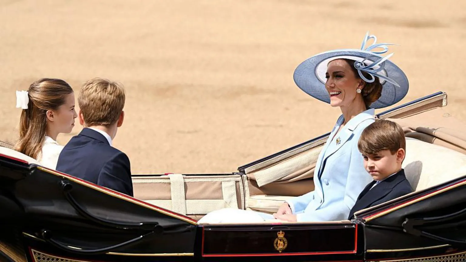 Princess Catherine sits in a carriage with a tan interior with Prince Louis, who sits next to her, and Princess Charlotte and Prince George, who sit opposite her. She is smiling and wearing a pale blue jacket with a pale blue hat. Louis and George are wearing dark suits and Charlotte is in white, with a white bow in her hair.