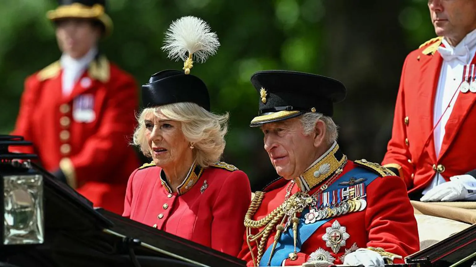 The King and Queen riding in a carriage. He is wearing the uniform of the Grenadier Guards as well as a number of medals. Queen Camilla is wearing a red, silk crepe, grenadier guards uniform dress and a black beret.