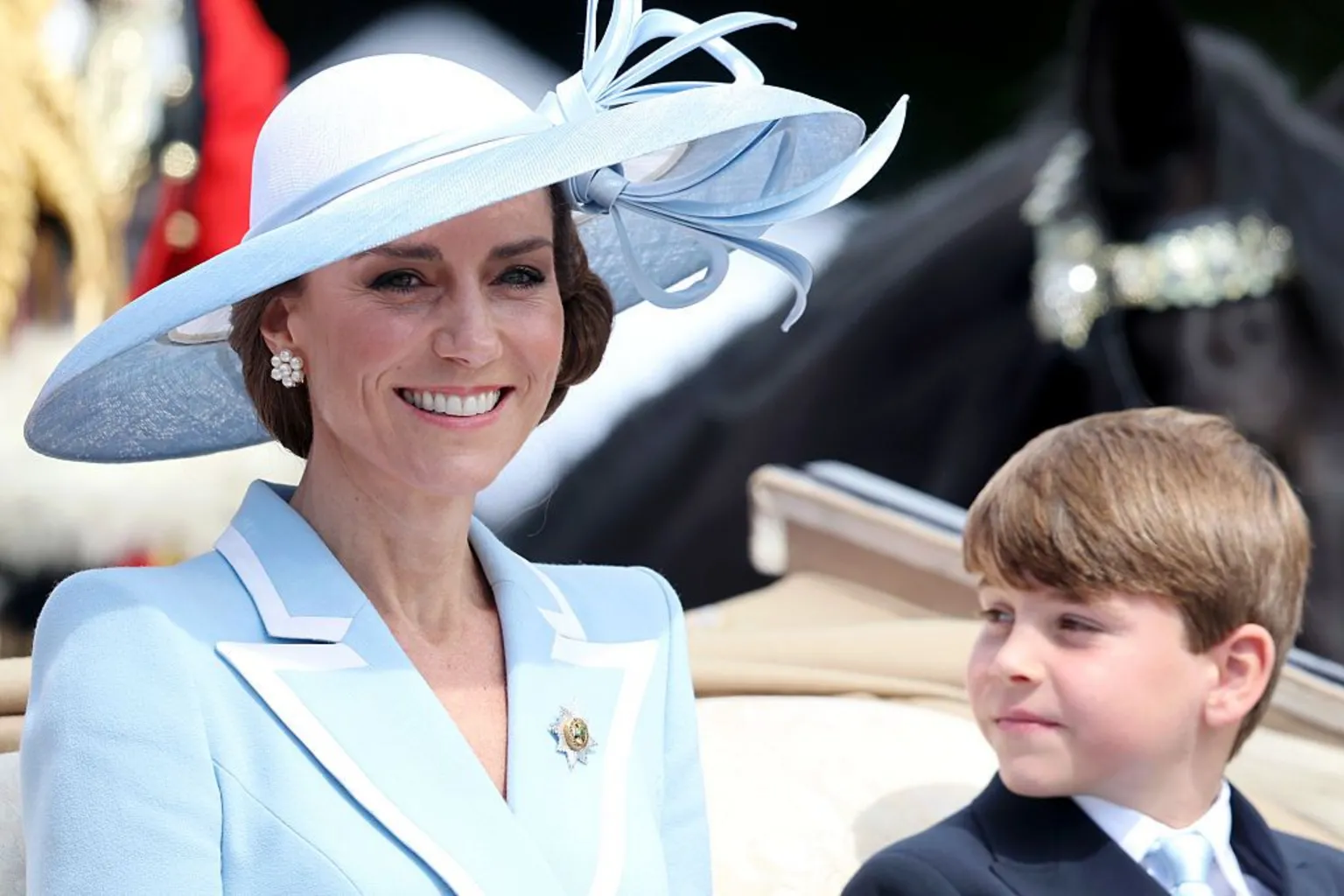 Princess Catherine smiles while sitting next to Prince Louis in a carriage, She is wearing a light blue wide-brimmed hat and a light blue jacket with white-trimmed lapels and a brooch. Louis is wearing a suit with a light blue tie.