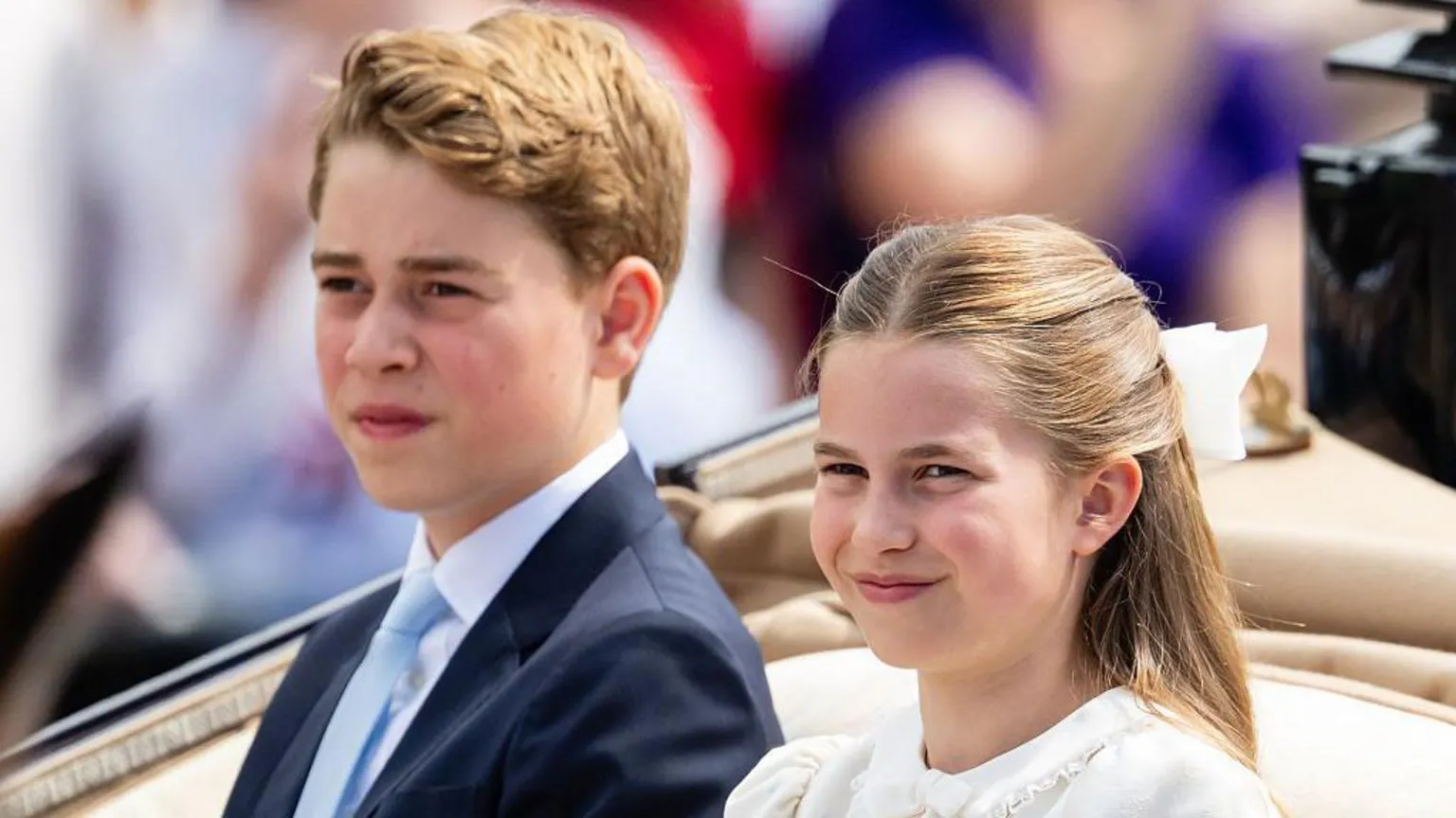 Princess Charlotte and Prince George ride in a carriage with a tan interior. She is wearing a white dress with a white bow in her hair and is smiling. He is wearing a dark suit with a white shirt and a light blue tie.
