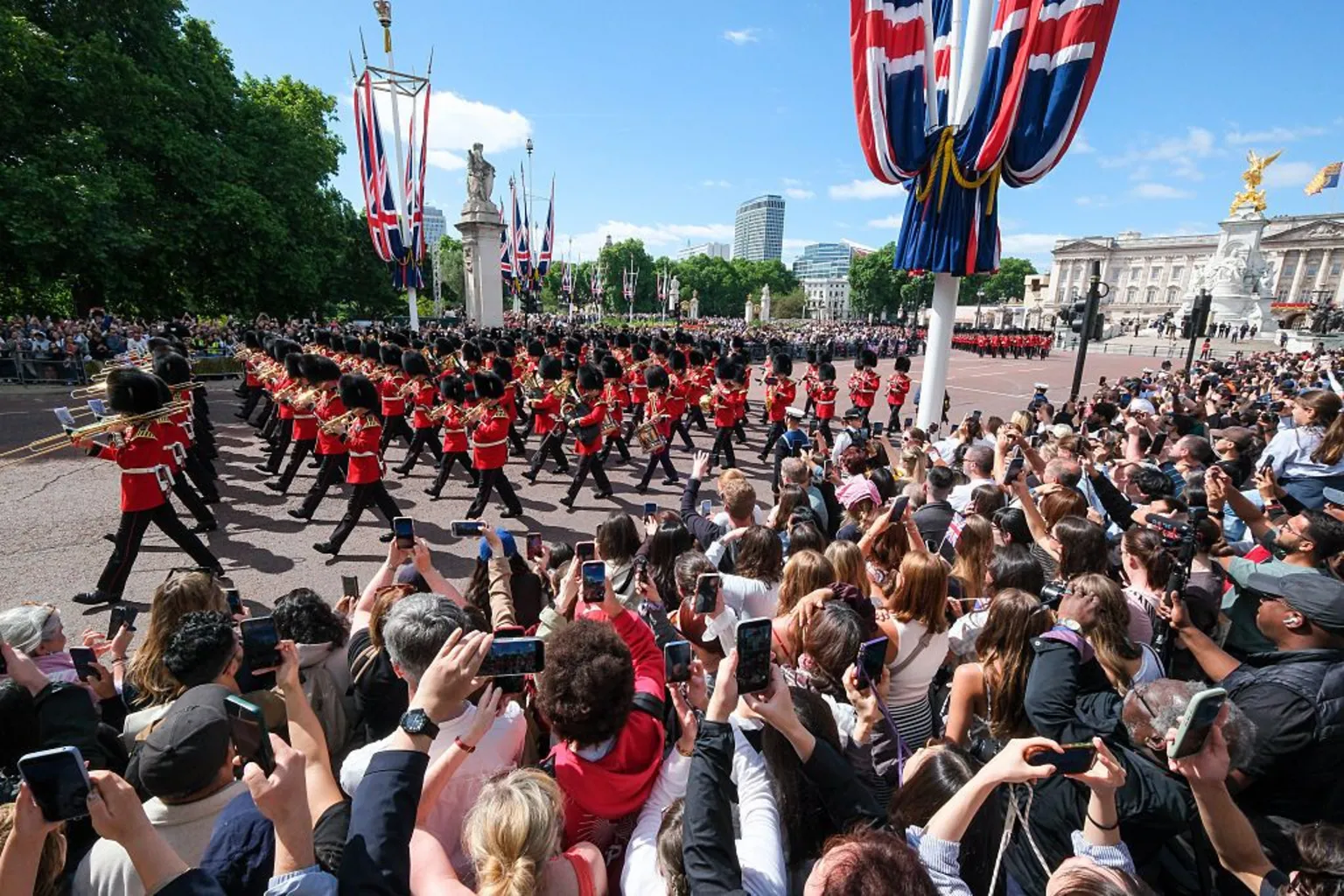 Crowds line both sides of the Mall and watch musicians from the Massed Bands of the Household Division parade from Buckingham Palace. Rows of onlookers stand in the sunshine, many with their mobile phones aloft. On the Mall, musicians in red, white and black uniform parade and play their instruments.