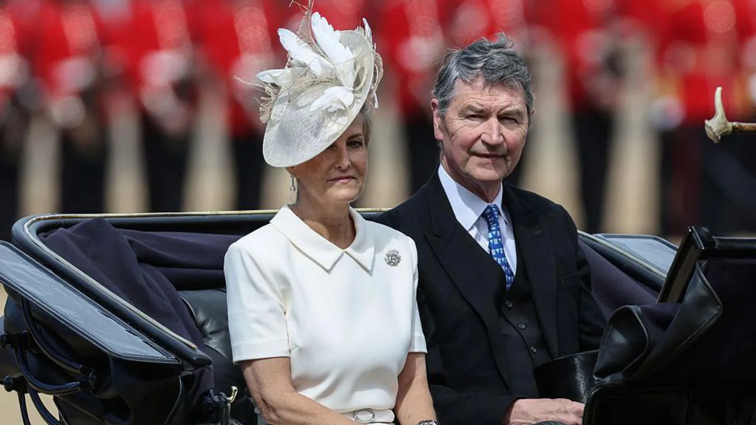 The Duchess of Edinburgh and Vice-Admiral Sir Tim Laurence ride in a black carriage. She is wearing a white dress with a collar and a brooch with a white hat. He is wearing a dark suit with a blue tie.