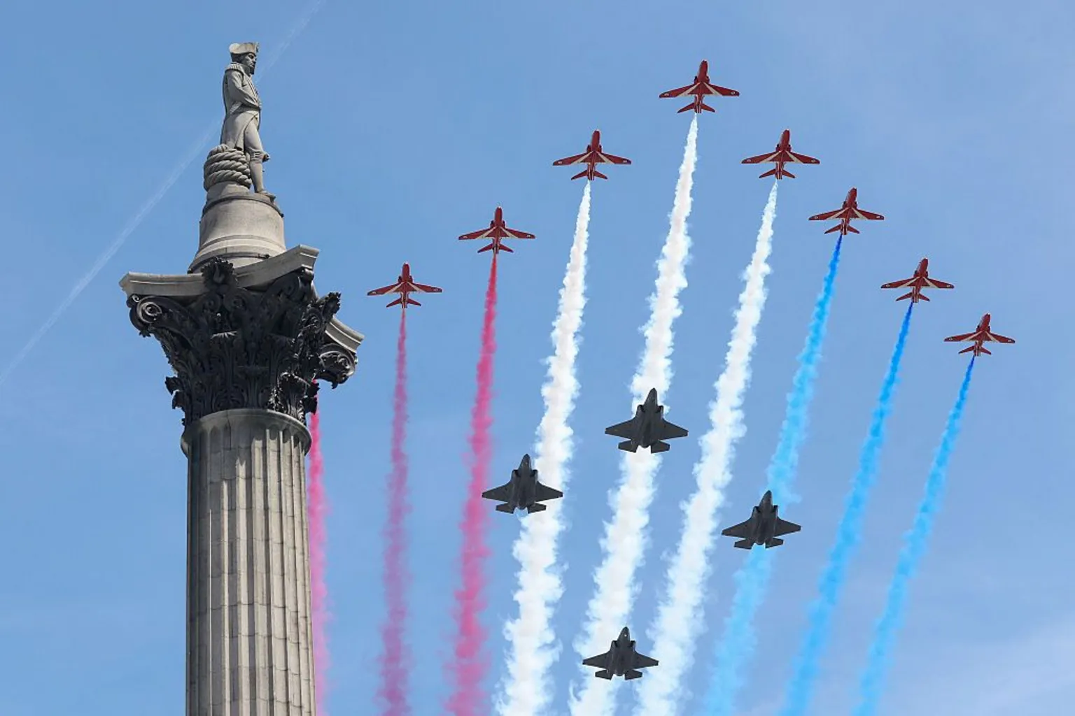 The Red Arrow take part in the flypast over Buckingham Palace. Twelve aircraft are pictured with plumes of red, white and blue clouds in their wake through a blue sky.