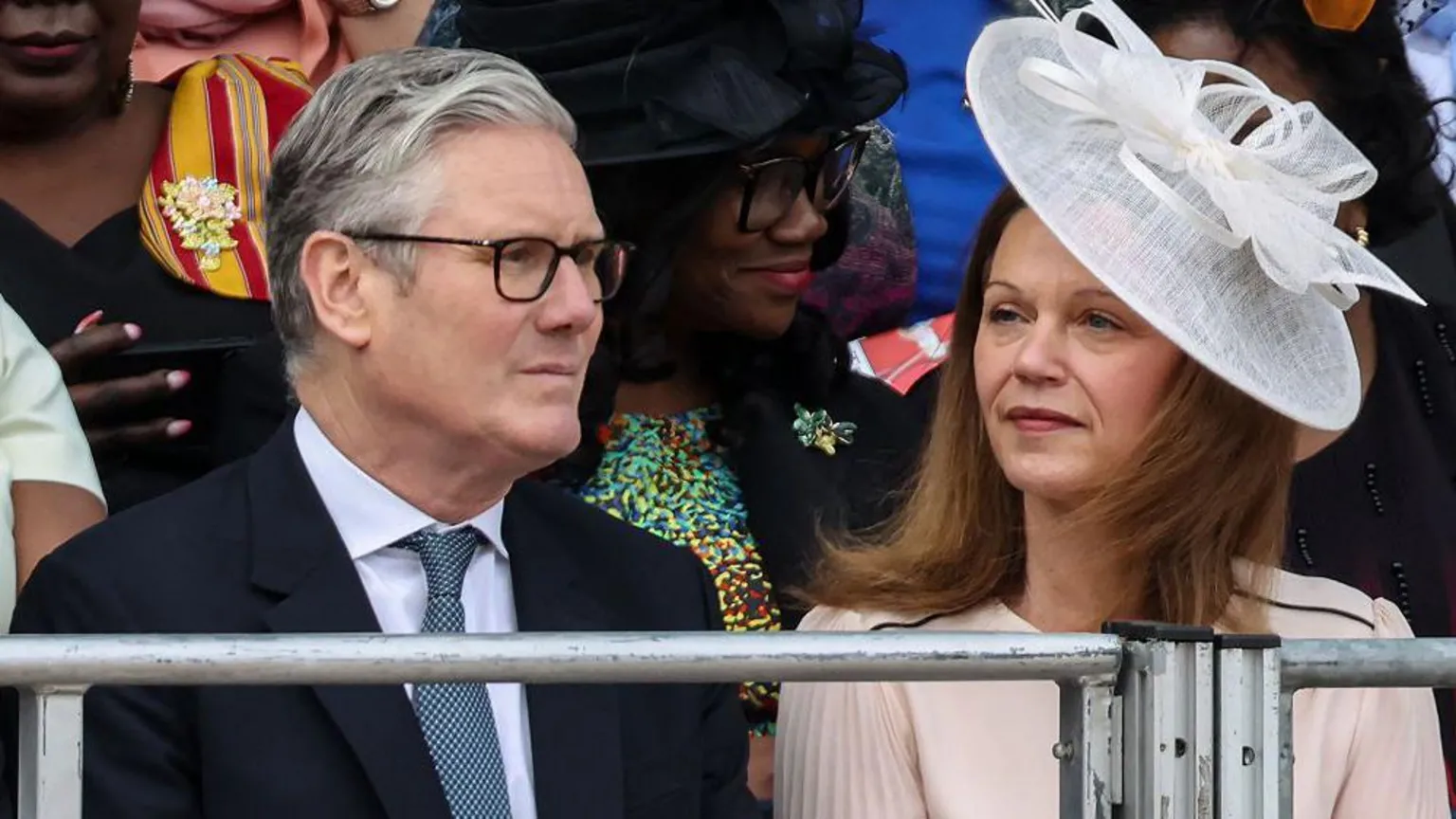 Sir Keir Starmer and Victoria Starmer sit to watch the parade. He is wearing a dark suit with a white shirt and a grey tie, and she wears a pale pink dress with a white hat.