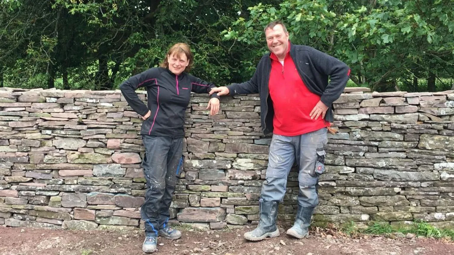 Christine Tallon Adrian Cole (right) and Christine Tallon (left) stood next to a stone wall. Behind the wall are several trees.