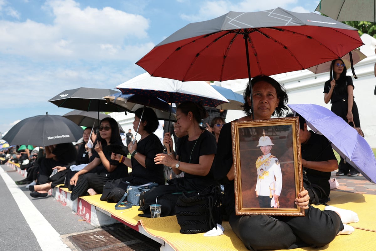 People shield themselves from the sun with umbrellas as they sit on a pavement, one holding a framed portrait of the princess
