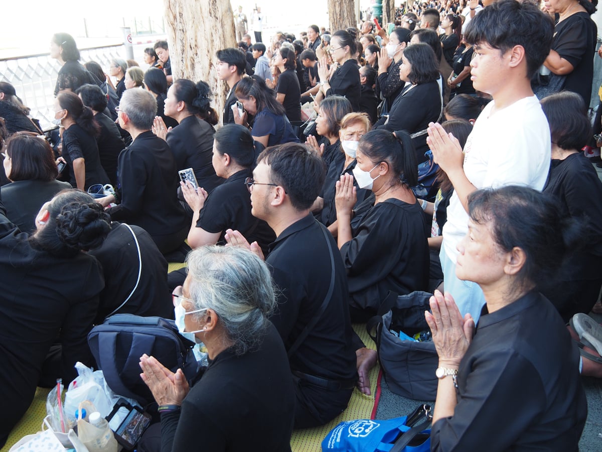A crowd of seated and standing mourners make a praying gesture with their hands as they look on
