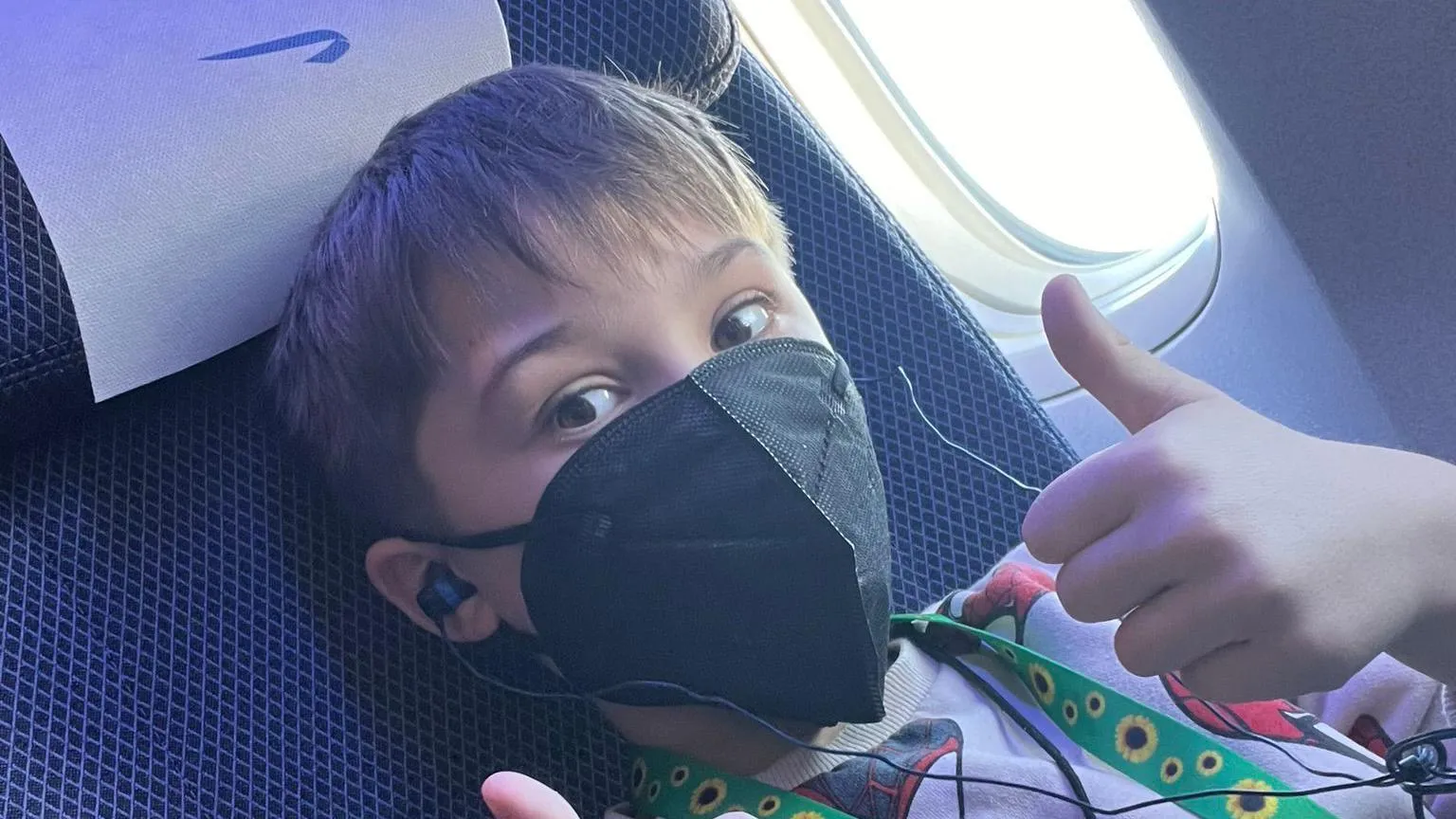Katie Hutt Yann Jennings, a 10-year-old boy, sat in an airplane seat wearing a black face mask, headphones, and a green lanyard with sunflowers on it. Giving thumbs up to the camera.