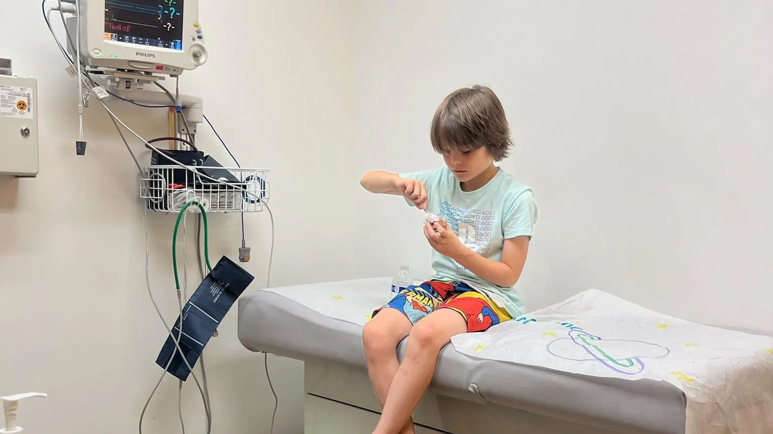 Katie Hutt A boy in a medical room wearing shorts and a t-shirt while eating pudding on a bed.